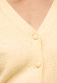 Mekana žuta pletena veste s tkanim gumbima preko golog tijela, prikazujući V-izrez i teksturirane detalje tkanine.