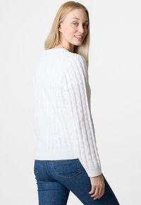 White knitted sweater with a textured, cable pattern, featuring a round neckline and ribbed cuff details. Worn with blue jeans.