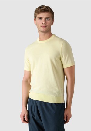 Joven con cabello corto que lleva un suéter de manga corta de color amarillo pálido y pantalones azul oscuro, de pie contra un fondo claro y liso.