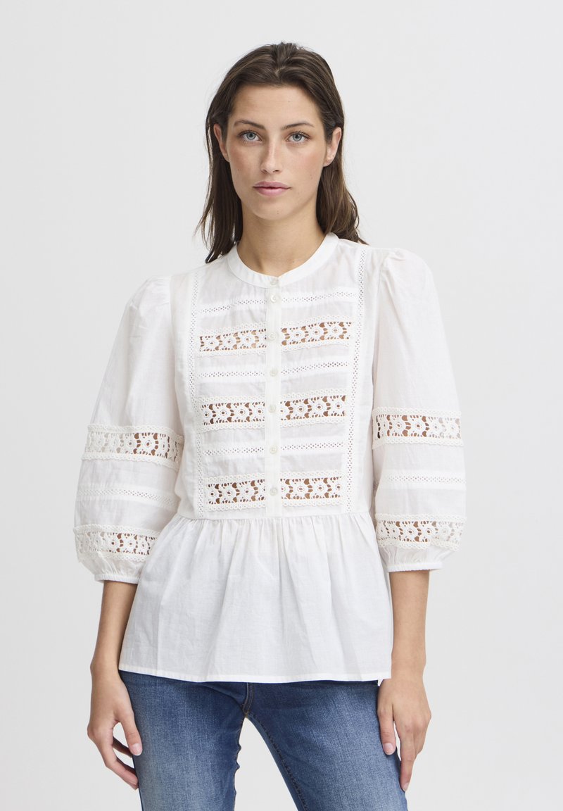 Blusa bianca con maniche a sbuffo, dettagli in pizzo, chiusura con bottoni e taglio peplo. Tessuto di cotone morbido con accenti testurizzati.