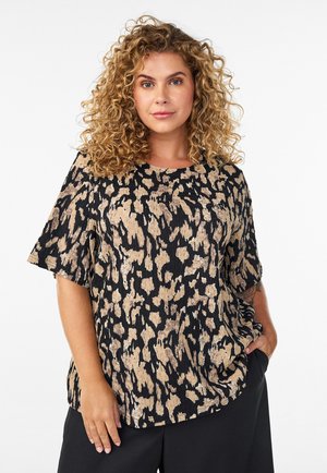 Femme aux cheveux bouclés portant un haut à manches courtes ample à imprimé animalier et un pantalon noir, debout avec une main dans la poche, fond neutre.