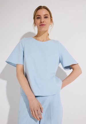 Frau mit blonden Haaren, die ein hellblaues Kurzarm-Oberteil und passende Hosen trägt, steht vor einem einfarbigen hellen Hintergrund.