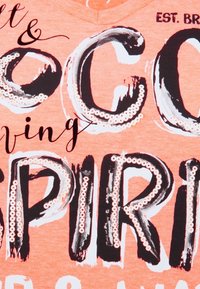 Camisa de algodón naranja con tipografía audaz en blanco y negro con acentos brillantes. El texto dice "Coco Living Spirit", con un diseño artístico de pinceladas.