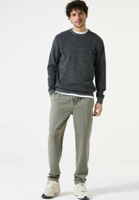 Grijze trui met een textuurpatroon, ronde hals en een klein logopatch, gecombineerd met lichtgroene, taps toelopende broek en witte sneakers.