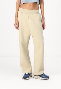 Bež široke trenirke iz mehke tkanine, z rebrastim pasom in belim napisom na strani. Kombinirane z modrimi športnimi čevlji.