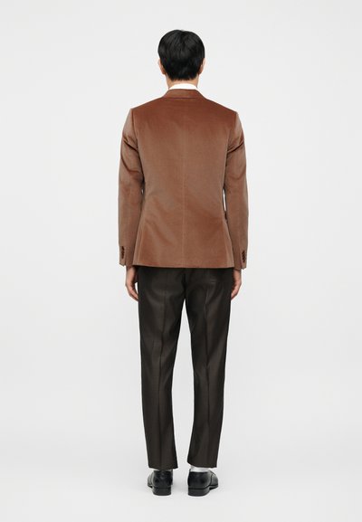 Καφέ σακάκι από κορδόνι με δομημένο σχέδιο, συνδυασμένο με σκούρο καρό παντελόνι και μαύρα παπούτσια. Ομαλή υφή, καθαρές γραμμές, ελάχιστα στολίδια.