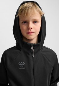 Svart huvtröja med dragkedja, slät tygtextur och en reflekterande logotyp. Designad med en figurnära passform och ergonomiska sömmar.