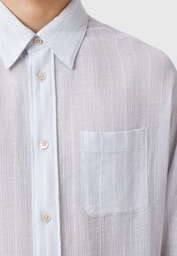 Vaaleansininen paitapusero, jossa on teksturoitu pystyraitakuosi, rintatasku ja vaaleat napit.