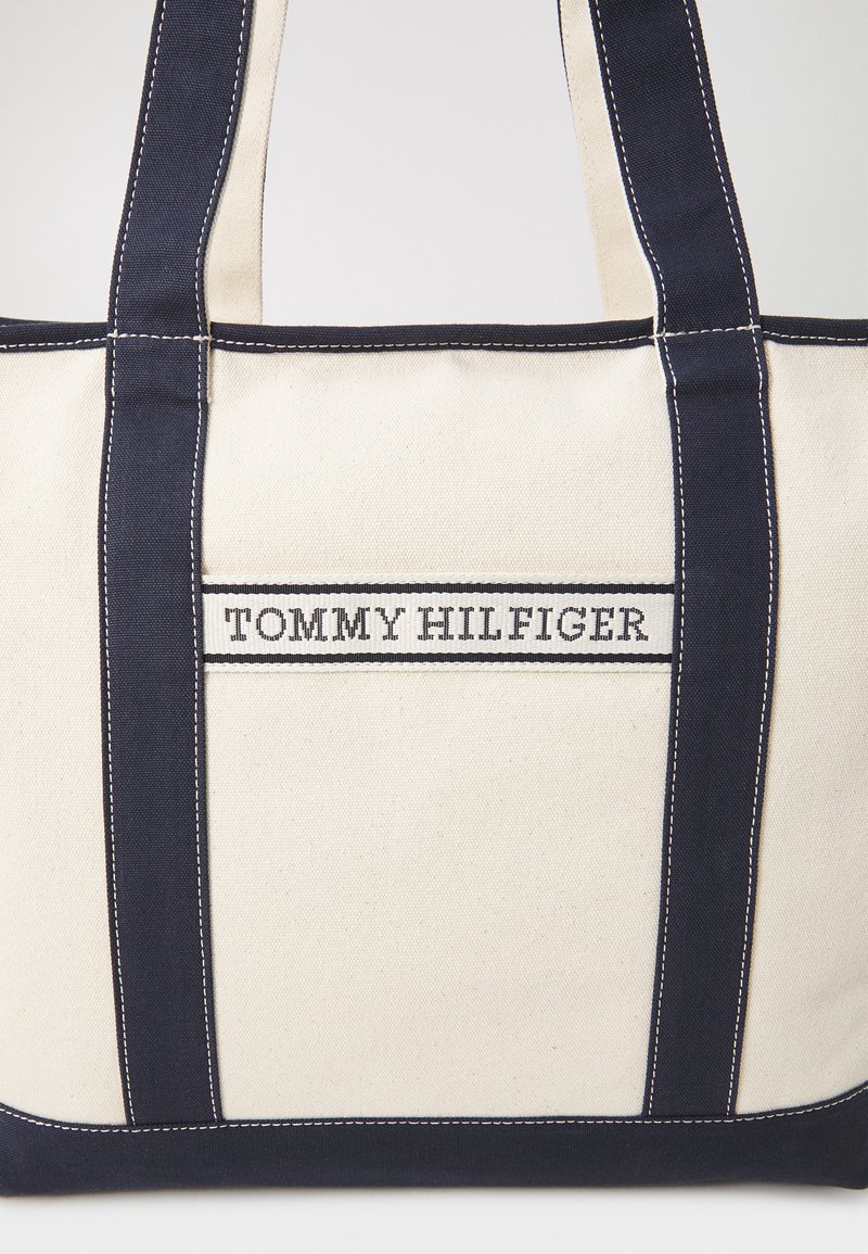 Torba płócienna w kolorze kremowym i granatowym, z solidnymi uchwytami, kontrastowym przeszyciem oraz naszywką z logo z napisem "TOMMY HILFIGER" na przodzie.
