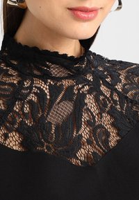 Frau trägt ein schwarzes, transparentes Spitzenoberteil mit hohem Kragen und Blumenmustern sowie große silberne Creolen, die Lippen leicht geöffnet.