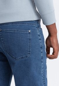 Persona con jeans de mezclilla azul y detalle en el bolsillo trasero, con una manga de suéter gris claro, relajada a un lado.