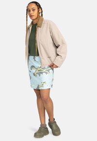 Beige zip-up jack met een bruine kraag, groene top, blauw met bloemenprint rok en groene klompen. Heeft bloemenpatronen en een gestructureerde stof.