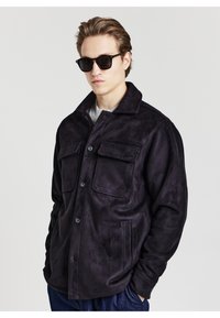 Lila Wildlederhemdjacke mit Kragen, zwei Brusttaschen und knöpfbarem Vorderteil. Modell trägt schwarze Sonnenbrille und graues Oberteil.
