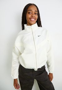Jauna sieviete ar gariem pītām matiem, tērpusies baltā rāvējslēdzēju jakā ar Nike logo un tumšiem rakstainiem biksēm, smaida un stāv pretī baltai fona.