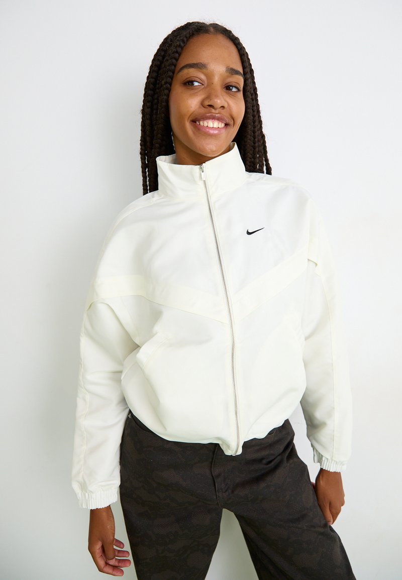 Young woman with long braids wearing white zip-up jacket with Nike logo and dark patterned pants, smiling and standing against white background.