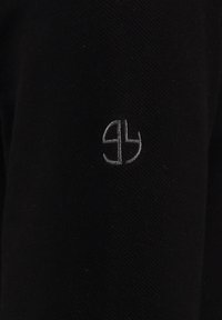 Prenda negra con una tela texturizada, adornada con un pequeño logo bordado en gris. El diseño es minimalista y moderno.