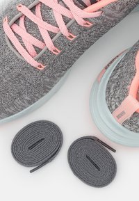 Chaussures de sport en tricot gris avec des lacets roses, présentées avec deux lacets gris supplémentaires enroulés sur un fond blanc.