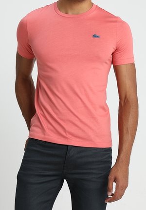 Homme portant un T-shirt ajusté à manches courtes couleur corail et un pantalon foncé, debout avec une main dans la poche devant un fond uni.