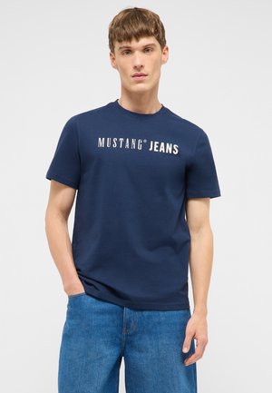 Junger Mann mit kurzen Haaren, der ein marineblaues "Mustang Jeans"-T-Shirt und blaue Jeans trägt, steht mit einer Hand in der Tasche.