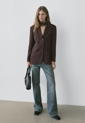 Kobieta w ciemnobrązowej marynarce, niebieskich dżinsach z szerokimi nogawkami, czarnych butach i trzymająca czarną torebkę na jednolitym szarym tle.