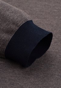 Strickpulloverbündchen aus geripptem marineblauem Material, kombiniert mit einem strukturierten braunen Stoff, der ein dezentes Karomuster aufweist.