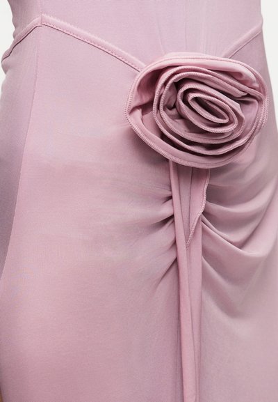 Κοντινό πλάνο ενός ανοιχτόροζ φορέματος με λεπτομέρεια διπλωμένου υφάσματος σε σχήμα τριαντάφυλλου στη μέση, δημιουργώντας απαλά ριγέ πτυχώσεις.