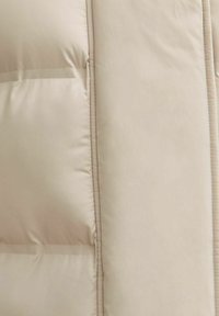 Chaqueta acolchada beige con una textura suave, que presenta costuras horizontales, dos bolsillos laterales y cierre de cremallera.