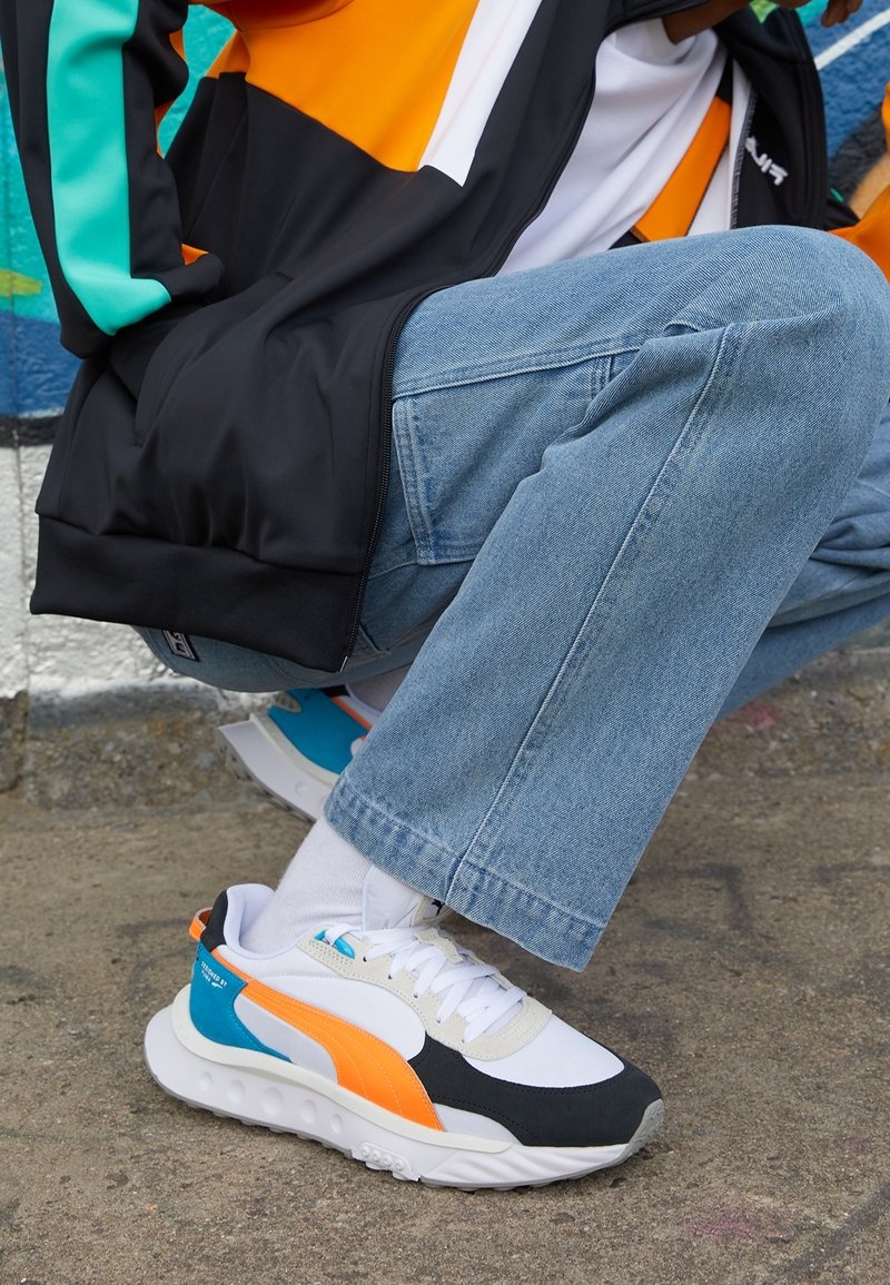 Sneakers con parte superiore bianca, punta nera, accenti arancioni e teal, suola texturizzata e lacci; posizionati su un abbigliamento casual in denim.