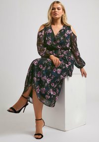 Blommig midi-klänning i genomskinlig svart tyg med lila och gröna blommor, design med kalla axlar, långa ärmar och en högt slits.