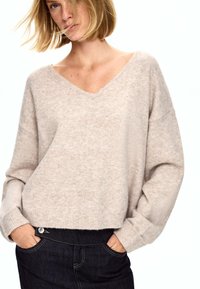 Svetlo bež, prevelik pulover iz mehkega pletenega materiala z V-izrezom in padajočimi rameni, v kombinaciji s temno modrimi jeans hlačami.