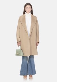 Manteau long en laine beige avec un large col, associé à un col roulé blanc, un jean évasé bleu, et un sac à main vert menthe. Chaussures couleur chair.