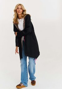 Černá kapucová bunda s podšívkou v leopardím vzoru, dlouhými rukávy, zipovým zapínáním a růžovými akcenty. Vydaná se světle modrými zúženými džíny a hnědými botami.