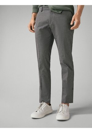 Homme portant un pantalon slim gris et des baskets blanches, debout devant un fond gris uni.