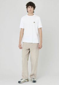 T-shirt en coton blanc avec un petit logo jaune sur la poitrine gauche, associé à un pantalon large beige texturé et des baskets multicolores.