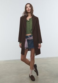 Brun ullkappa över en grön kortärmad cardigan, denimkjol med randig midja, och bruna loafers med ljusa strumpor.