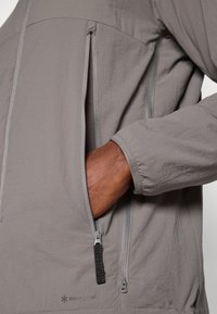 Veste légère grise en tissu doux, dotée d'une poche zippée et de poignets élastiques ; le logo sur le bas avant apporte un détail subtil.