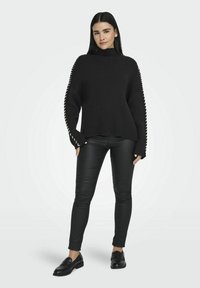 Maglione nero a coste con collo alto e dettagli in pizzo sulle maniche, abbinato a pantaloni neri e mocassini.