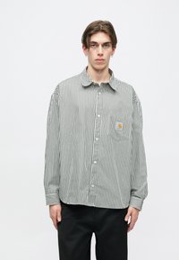 Camisa de botones a rayas en blanco y negro, de corte oversize, con un bolsillo en el pecho y una pequeña etiqueta de logo. Mangas largas con detalles en los puños.