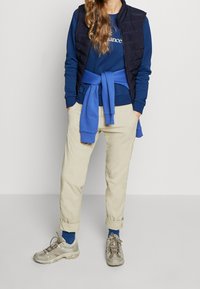 Modrá mikina s bílým textem, námořnická prošívaná vesta, béžové zrolované kalhoty a šedé turistické boty s modrými ponožkami.