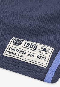 Marineblaue Stoff mit gerippter Textur, versehen mit einem weißen Etikett, einem Vintage-Logo und blauen Akzentstreifen. Das Etikett enthält "1908" und "Converse Ath. Dept."