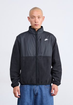 Mann mit glattem Kopf, der eine schwarze Nike-Fleecejacke und eine blaue Jeans trägt, steht vor einem schlichten weißen Hintergrund.