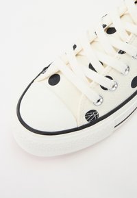 Trampki z płótna w czarne kropki na kremowym tle, z białym gumowym czubkiem, czarną lamówką i beżowymi sznurowadłami.