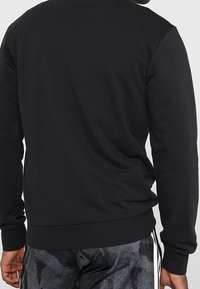 Uomo che indossa una felpa a maniche lunghe nera e pantaloni corti sportivi a righe nere e grigie, visto da dietro su uno sfondo semplice.