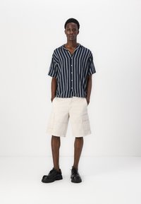Chemise à rayures marine à manches courtes avec un devant à bouton, associée à un short cargo beige clair et des chaussures noires à lacets.