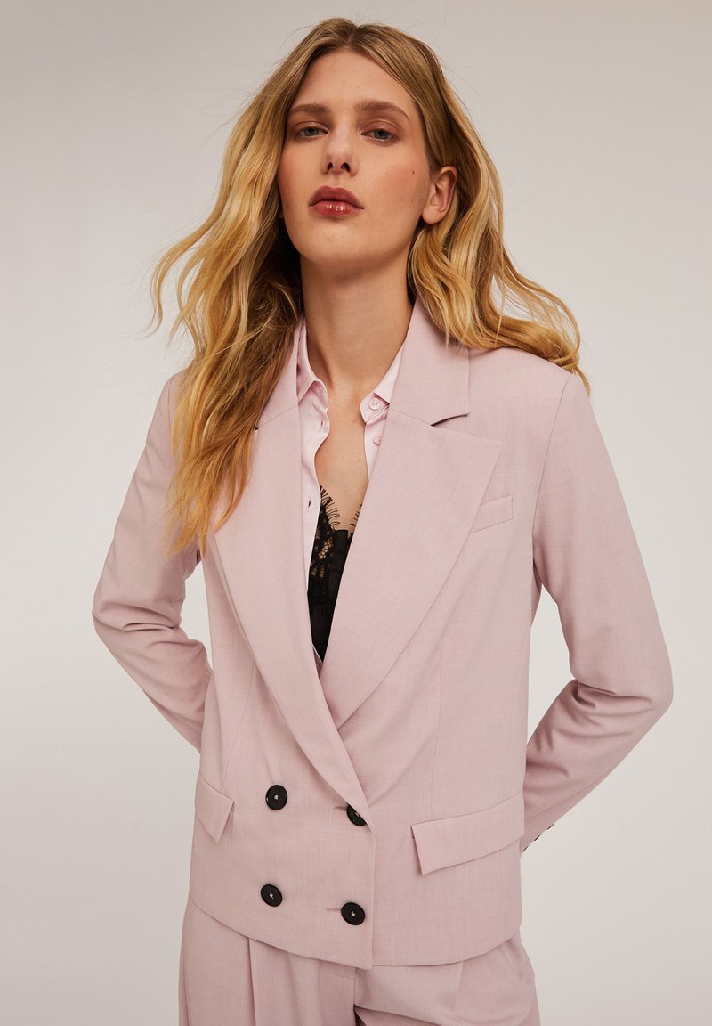 Blazer double boutonnage rose clair avec des boutons noirs, revers en pointe et poches latérales. Porté sur un haut en dentelle noir et une chemise rose clair.