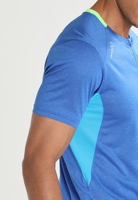Camisa deportiva azul de mangas cortas. Presenta un cuello verde y paneles laterales de azul claro. Está hecha de una tela suave y ligera.