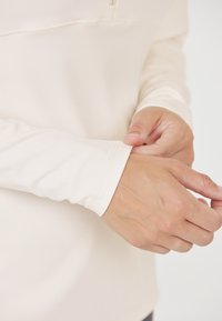 Leichtes Langarmoberteil in Creme, aus glattem Material gefertigt, mit einer Reißverschlusstasche und enganliegenden Bündchen mit dezenten Stickdetails.