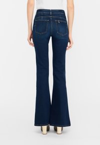 Jean en denim bleu foncé, coupe slim avec jambes évasées, porté par une personne debout, vue de dos sur fond blanc uni.