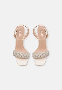 Beige hælhøye sandaler med en flettefølt metallisk stropp og firkantet tådesign. Har tynne ankelstropper og glatt tekstur.