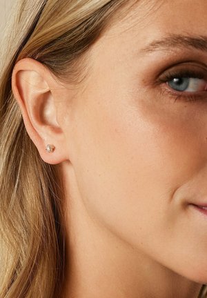 Das linke Ohr einer Frau mit einem kleinen goldenen Stecker, der einen einzelnen klaren Edelstein zeigt. Zu sehen sind hellbraune Haare und ein blauer Augen.